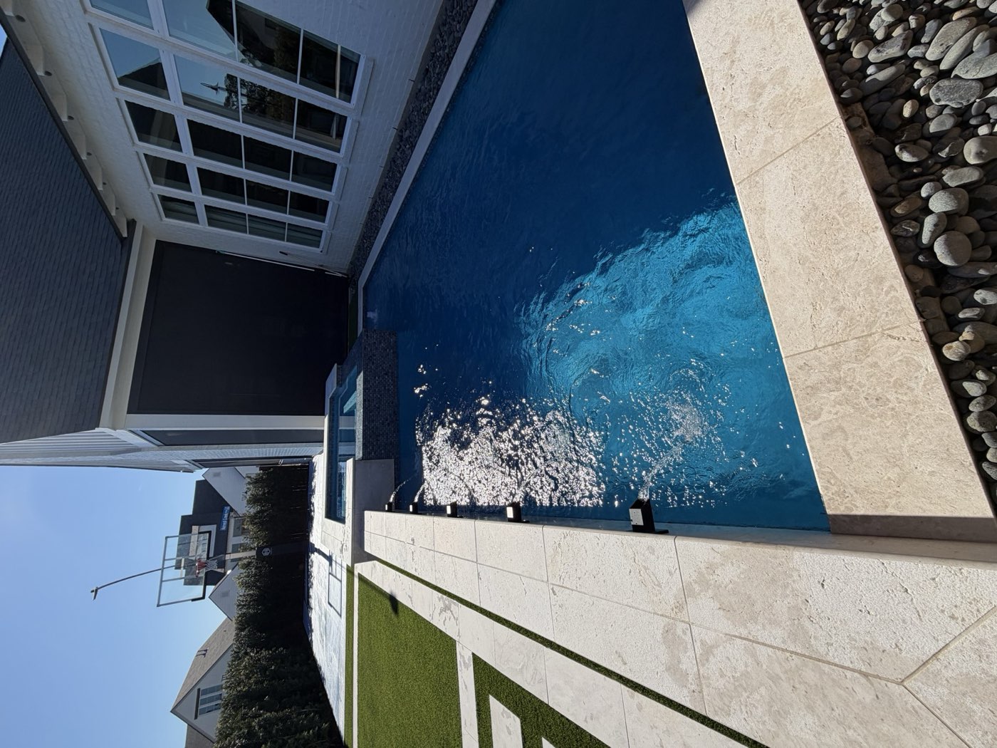 Modern pool with spa and turf beside white home with basketball court